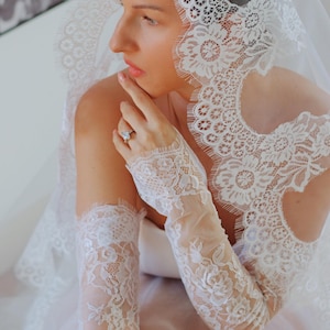 May include: White lace bridal gloves with a delicate floral pattern. The gloves are long and extend to the elbow. The gloves are made of a sheer fabric and have a scalloped edge.