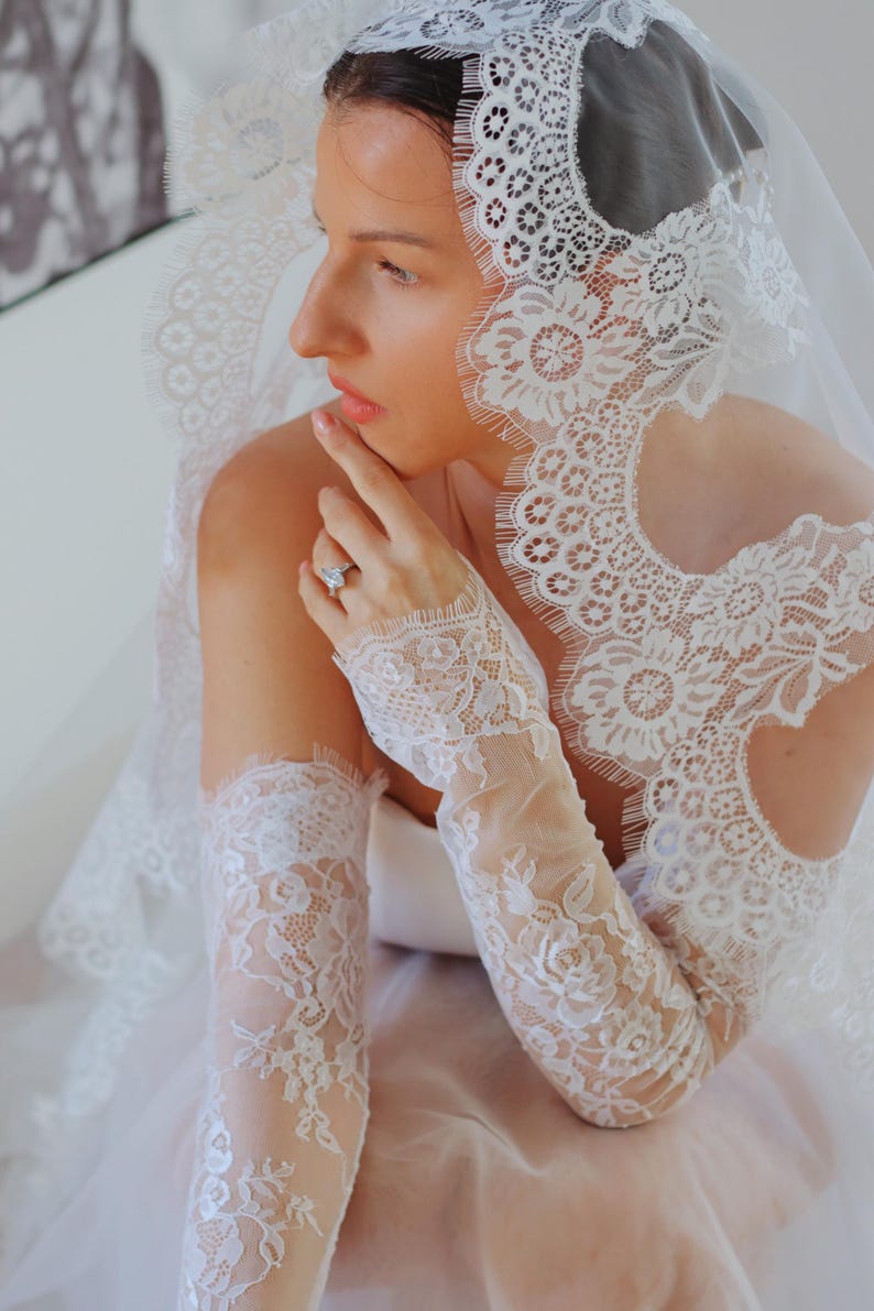 May include: White lace bridal gloves with a delicate floral pattern. The gloves are long and extend to the elbow. The gloves are made of a sheer fabric and have a scalloped edge.