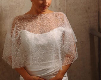 Könnte beinhalten: Ein weißes Brautkleid mit einem durchsichtigen, perlenbesetzten Umhang. Der Umhang ist mit kleinen, glitzernden Perlen verziert, die einen zarten, schimmernden Effekt erzeugen. Das Kleid hat ein schlichtes, elegantes Design mit einem figurbetonten Oberteil und einem fließenden Rock. Der Gesamteindruck ist anspruchsvoll und romantisch.