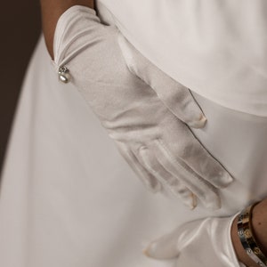 May include: A pair of white satin gloves with pearl accents. The gloves are worn on the hands of a person wearing a white dress and a gold bracelet.