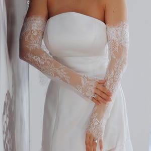 May include: A white strapless wedding dress with a tulle skirt and lace sleeves. The bride is wearing a ring on her finger.