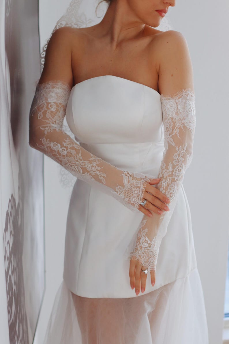 May include: A white strapless wedding dress with a tulle skirt and lace sleeves. The bride is wearing a ring on her finger.