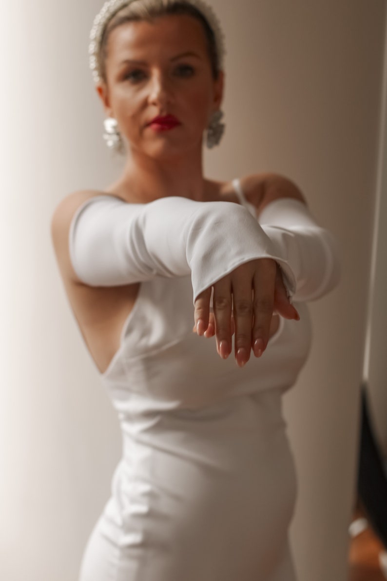 May include: A white satin dress with matching long gloves. The dress has a simple, elegant design, and the gloves extend to the elbows. The model is wearing a pearl headband and earrings, creating a classic, sophisticated look.