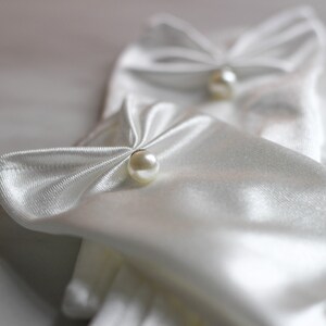 May include: A pair of white satin gloves with pearl embellishments. The gloves are folded and the pearls are visible on the top of the gloves.