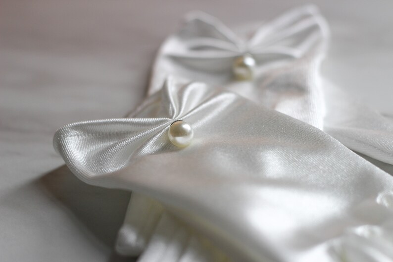 May include: A pair of white satin gloves with pearl embellishments. The gloves are folded and the pearls are visible on the top of the gloves.