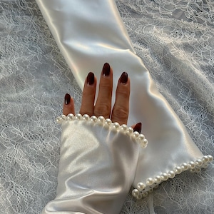 May include: White satin gloves with a pearl trim. The gloves are long and reach to the elbow. The pearls are small and evenly spaced.