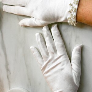May include: A pair of white satin gloves with pearl trim. The gloves are shown on a white marble surface.