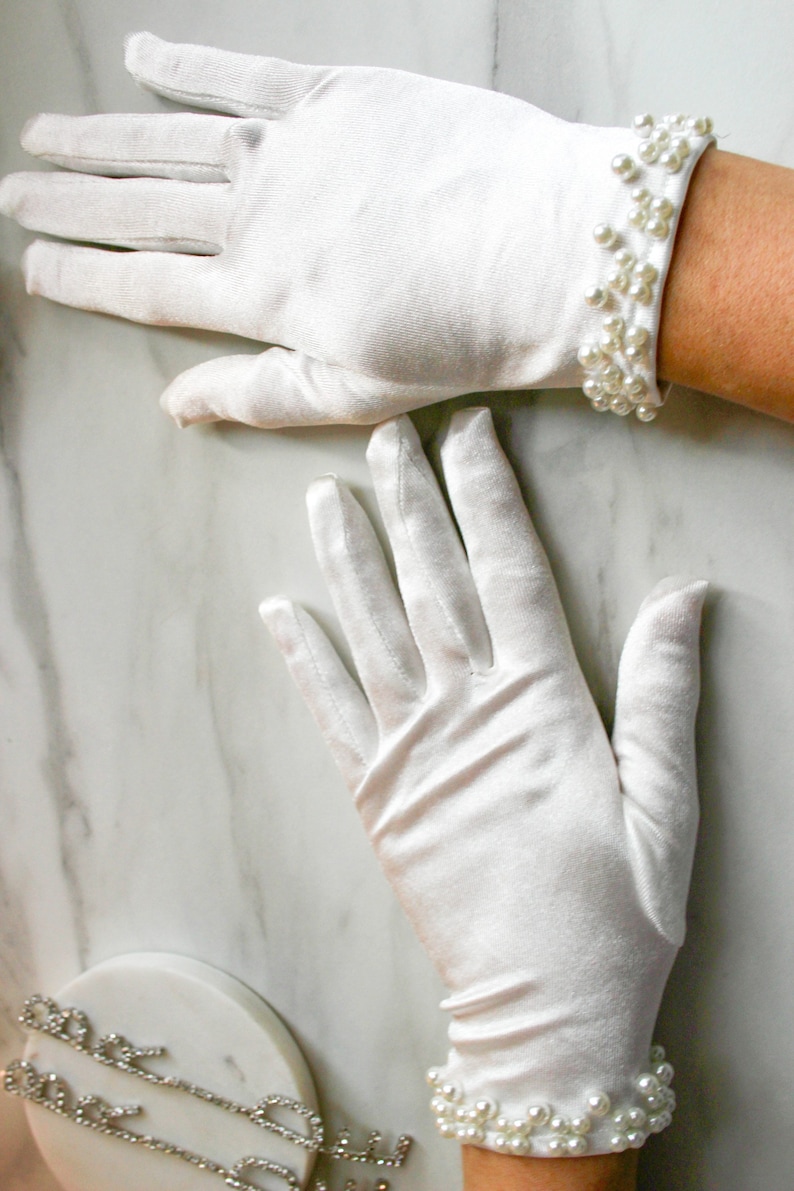 May include: A pair of white satin gloves with pearl trim. The gloves are shown on a white marble surface.