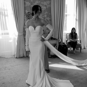 May include: A strapless white wedding gown with a sweetheart neckline and a long, flowing train. The dress is paired with long, matching gloves. The bride is standing in a room with large windows and curtains.