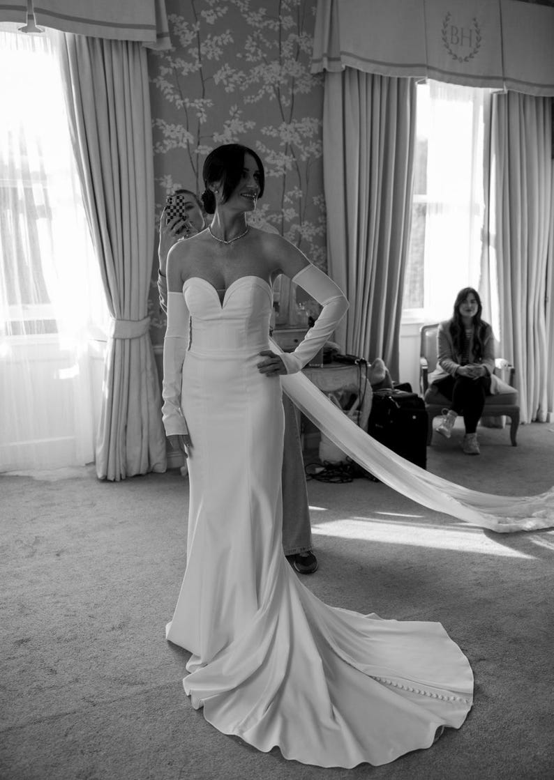 May include: A strapless white wedding gown with a sweetheart neckline and a long, flowing train. The dress is paired with long, matching gloves. The bride is standing in a room with large windows and curtains.