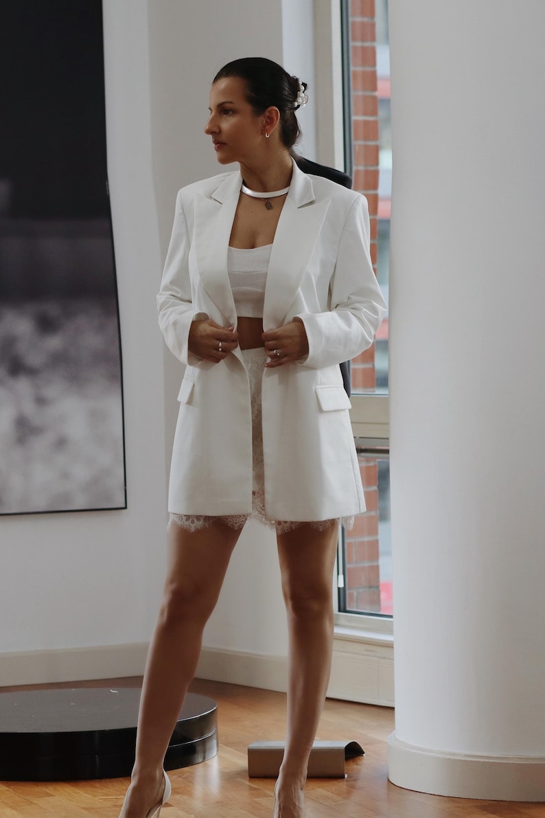 May include: A white blazer with a white crop top and a white lace skirt.