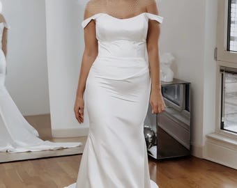 Könnte beinhalten: Weißes schulterfreies Brautkleid mit Meerjungfrau-Silhouette. Das Kleid hat ein eng anliegendes Oberteil und einen fließenden Rock. Die Braut trägt einen Schleier. Der Raum hat einen Spiegel und Holzboden.