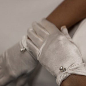 May include: A pair of white satin gloves with pearl embellishments. The gloves are worn on a person's hands.