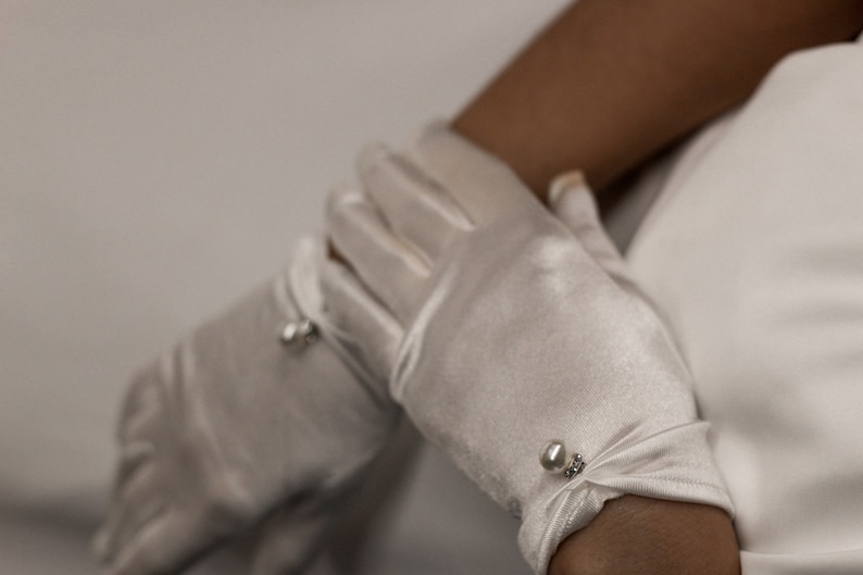 May include: A pair of white satin gloves with pearl embellishments. The gloves are worn on a person's hands.