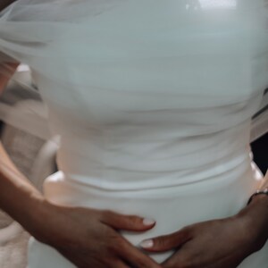 May include: A white wedding dress with a one-shoulder design and a flowing tulle overlay. The dress is fitted and hugs the body.