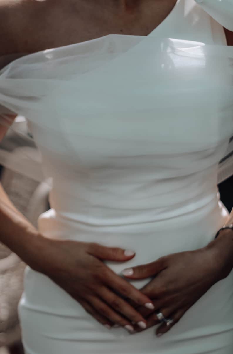 May include: A white wedding dress with a one-shoulder design and a flowing tulle overlay. The dress is fitted and hugs the body.