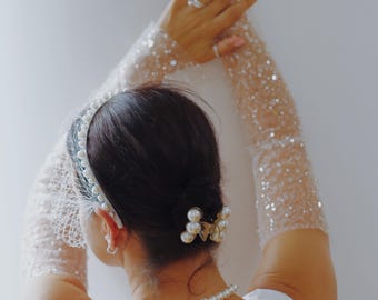 May include: A bride is seen from behind, wearing a white dress, pearl necklace, and a pearl and lace headband. She has a pearl hair clip and sheer, sequined gloves. The image shows arms raised.