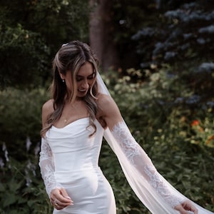 May include: A white floor-length gown with a cowl neckline and fitted bodice, featuring detachable sheer lace sleeves that extend into long flowing fabric panels, paired with a matching sheer veil attached to the back of the hair.