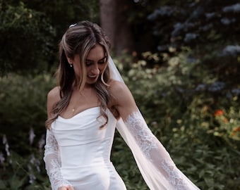 May include: A white floor-length gown with a cowl neckline and fitted bodice, featuring detachable sheer lace sleeves that extend into long flowing fabric panels, paired with a matching sheer veil attached to the back of the hair.