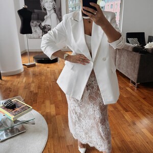 May include: A white blazer with a double breasted closure and gold buttons. The blazer is paired with a white lace skirt and white heels.