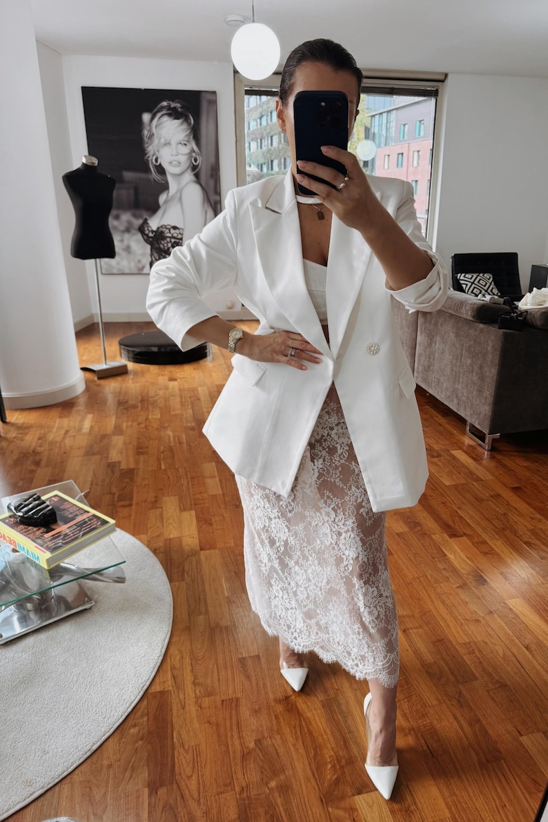 May include: A white blazer with a double breasted closure and gold buttons. The blazer is paired with a white lace skirt and white heels.