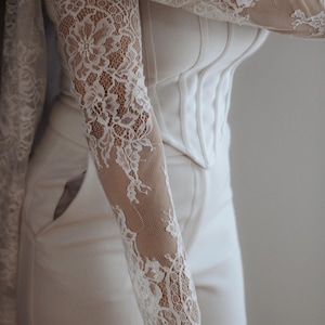 May include: A white corset-style top featuring long, sheer lace sleeves with intricate floral patterns and a scalloped edge at the wrists. The bodice is structured with vertical seams and paired with matching white trousers that have side pockets.