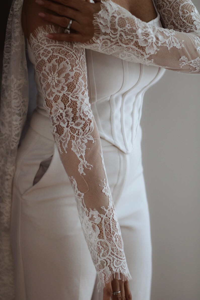 May include: A white corset-style top featuring long, sheer lace sleeves with intricate floral patterns and a scalloped edge at the wrists. The bodice is structured with vertical seams and paired with matching white trousers that have side pockets.