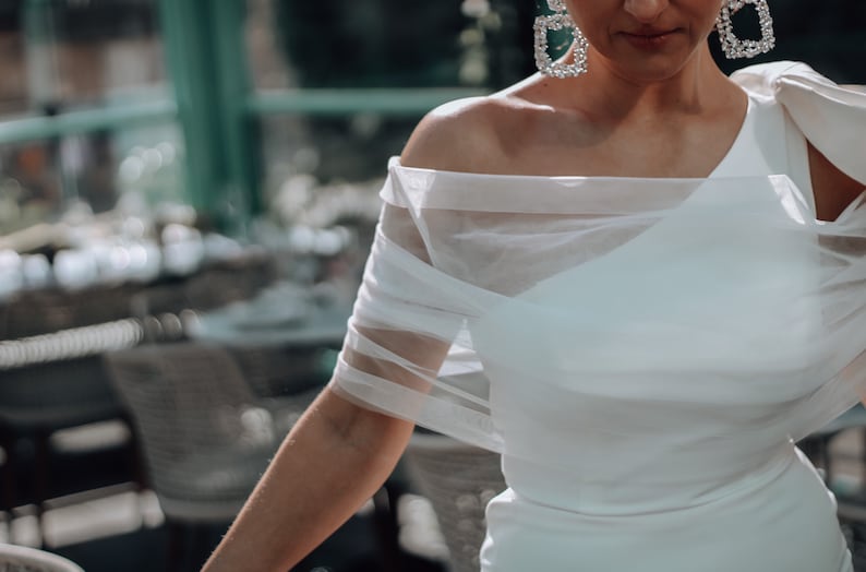 May include: A white off-the-shoulder dress with a sheer overlay. The dress has a simple, elegant design and is perfect for a special occasion.