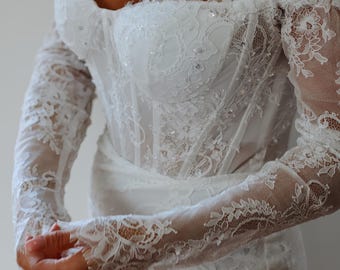 May include: A white, off-the-shoulder wedding dress with intricate lace detailing. The bodice is fitted and embellished with sequins, while the long sleeves and neckline feature delicate lace trim. The dress appears to be made of a soft, flowing fabric.