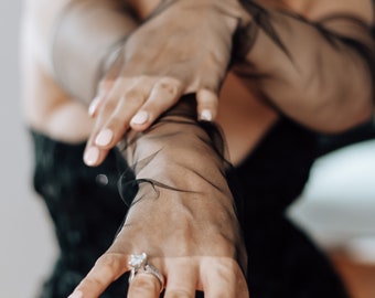 May include: A close-up of a hand wearing a black sheer glove with a diamond engagement ring on the ring finger.