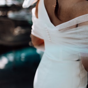May include: A white bridal gown with a sheer overlay and a low back. The gown has a fitted bodice and a flowing skirt.