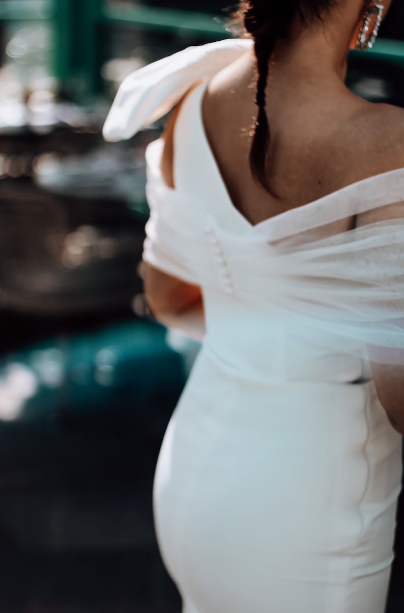 May include: A white bridal gown with a sheer overlay and a low back. The gown has a fitted bodice and a flowing skirt.