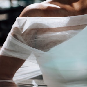 May include: A white sheer fabric overlay with a one-shoulder design. The fabric is draped over the shoulder and falls down the arm.