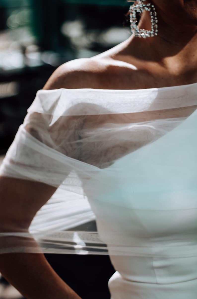 May include: A white sheer fabric overlay with a one-shoulder design. The fabric is draped over the shoulder and falls down the arm.