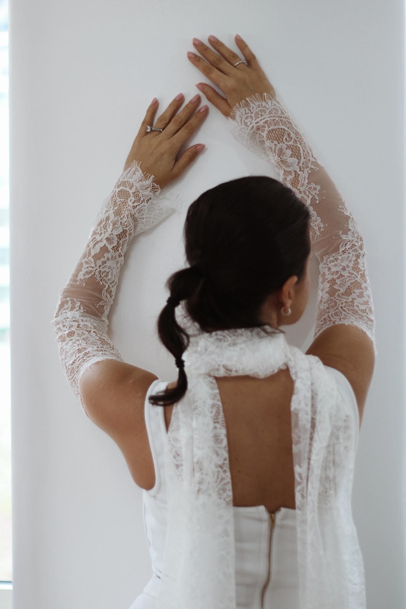 May include: A pair of white lace arm sleeves featuring an intricate floral pattern with scalloped edges that extend from the wrist to the upper arm, paired with a matching white lace scarf draped around the neck over a white sleeveless top.