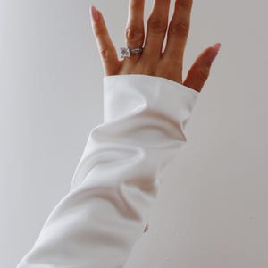 Pode incluir: Uma mão com unhas compridas e cor-de-rosa usando um anel de prata com uma pedra grande, quadrada e transparente. A mão emerge de uma camisa branca de manga comprida contra um fundo branco.