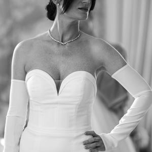 May include: A strapless white wedding dress with a sweetheart neckline and long, fitted gloves. A sparkling necklace adorns the neck. The dress is form-fitting and elegant, suitable for a formal event.