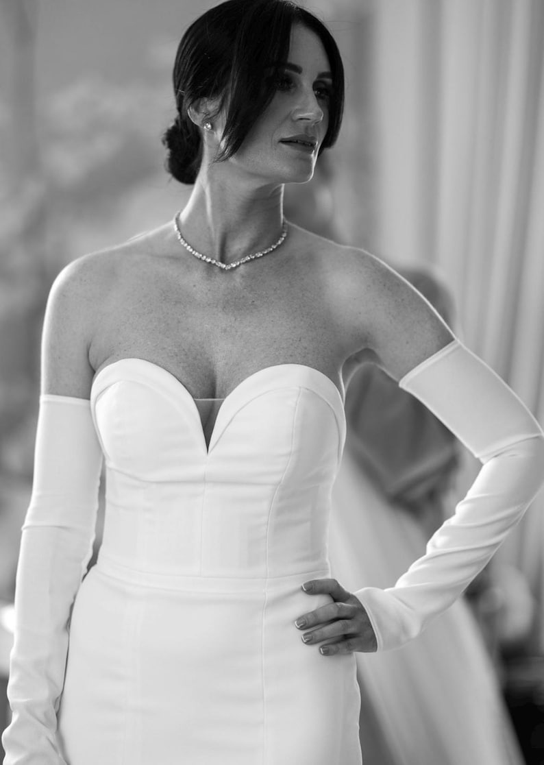 May include: A strapless white wedding dress with a sweetheart neckline and long, fitted gloves. A sparkling necklace adorns the neck. The dress is form-fitting and elegant, suitable for a formal event.