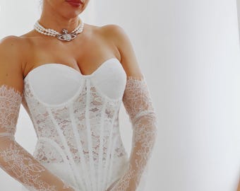 Puede incluir: Un top de corsé de encaje blanco sin tirantes y una falda a juego. El corsé presenta ballenas verticales y un escote corazón. Guantes de encaje y un collar de perlas completan el conjunto. Adecuado para una boda o evento especial.
