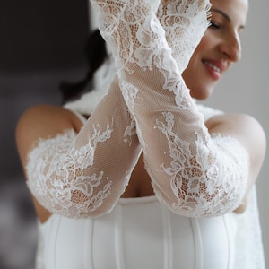 May include: A pair of white lace arm sleeves featuring an intricate floral pattern and a delicate scalloped edge at the wrist. The sleeves are made of a fine, semi-sheer mesh fabric that extends from the wrist up to the upper arm area.