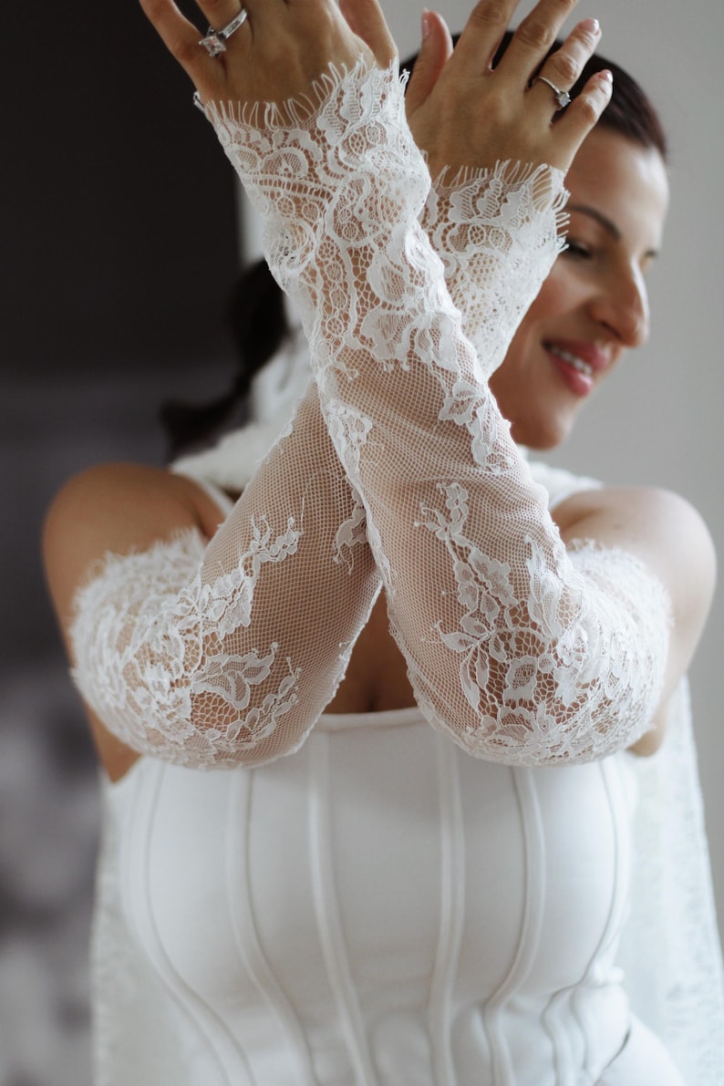 May include: A pair of white lace arm sleeves featuring an intricate floral pattern and a delicate scalloped edge at the wrist. The sleeves are made of a fine, semi-sheer mesh fabric that extends from the wrist up to the upper arm area.
