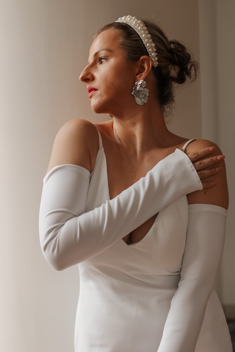 May include: A woman wearing a white dress with long sleeves and a pearl headband. The dress has a low neckline. Silver-colored earrings are visible. The woman is looking to the side.
