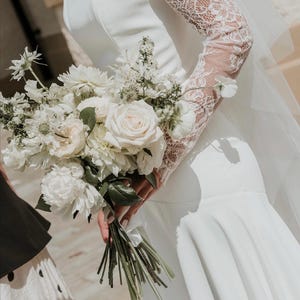May include: A strapless white gown featuring a fitted bodice and a flared skirt, paired with long, sheer lace sleeves. The garment is accompanied by a large, textured bouquet of white roses, peonies, and greenery, held by the side of the dress.
