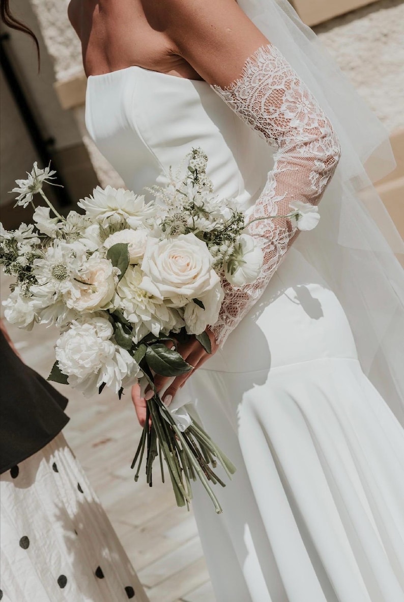 May include: A strapless white gown featuring a fitted bodice and a flared skirt, paired with long, sheer lace sleeves. The garment is accompanied by a large, textured bouquet of white roses, peonies, and greenery, held by the side of the dress.