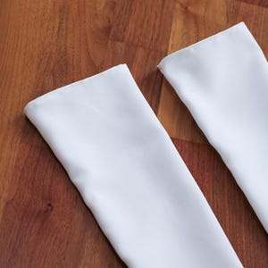 May include: Two white rectangular fabric pieces with folded edges, likely napkins or placemats, are arranged diagonally against a warm, brown wooden surface. The fabric appears smooth and clean, contrasting with the wood grain.