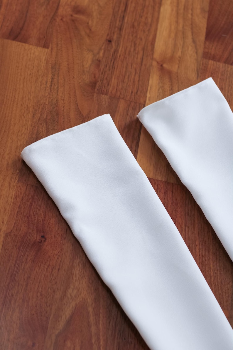 May include: Two white rectangular fabric pieces with folded edges, likely napkins or placemats, are arranged diagonally against a warm, brown wooden surface. The fabric appears smooth and clean, contrasting with the wood grain.
