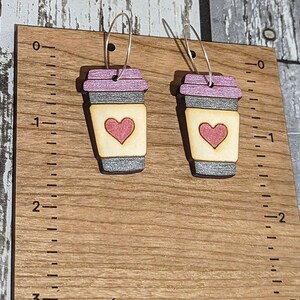 May include: A pair of wooden coffee cup earrings with a pink glitter finish and a pink heart design. The earrings are hanging from silver hoops.
