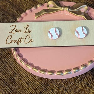 May include: A pair of white baseball-themed earrings with red stitching on a pink and gold display stand. The stand has a wooden sign that reads "Zoe Ly Craft Co."