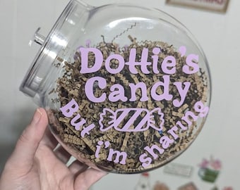 May include: A clear, round plastic jar with a slanted opening and a lid. The jar features light purple vinyl lettering that reads Dottie's Candy But I'm sharing, with a candy graphic in the center. The interior is filled with brown paper filler.