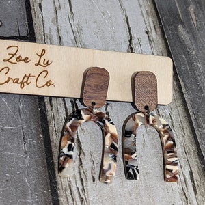 May include: A pair of brown and white patterned resin earrings with a wood accent. The earrings are shaped like a horseshoe and are hanging from a wooden display.